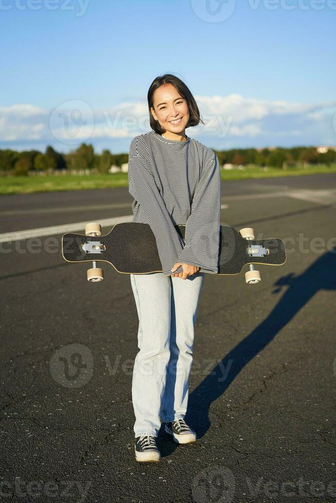 vertical tiro do skatista menina posando com Longboard, cruzeiro em esvaziar estrada dentro subúrbios. sorridente ásia mulher patinação em skate, segurando cruzador dentro mãos foto