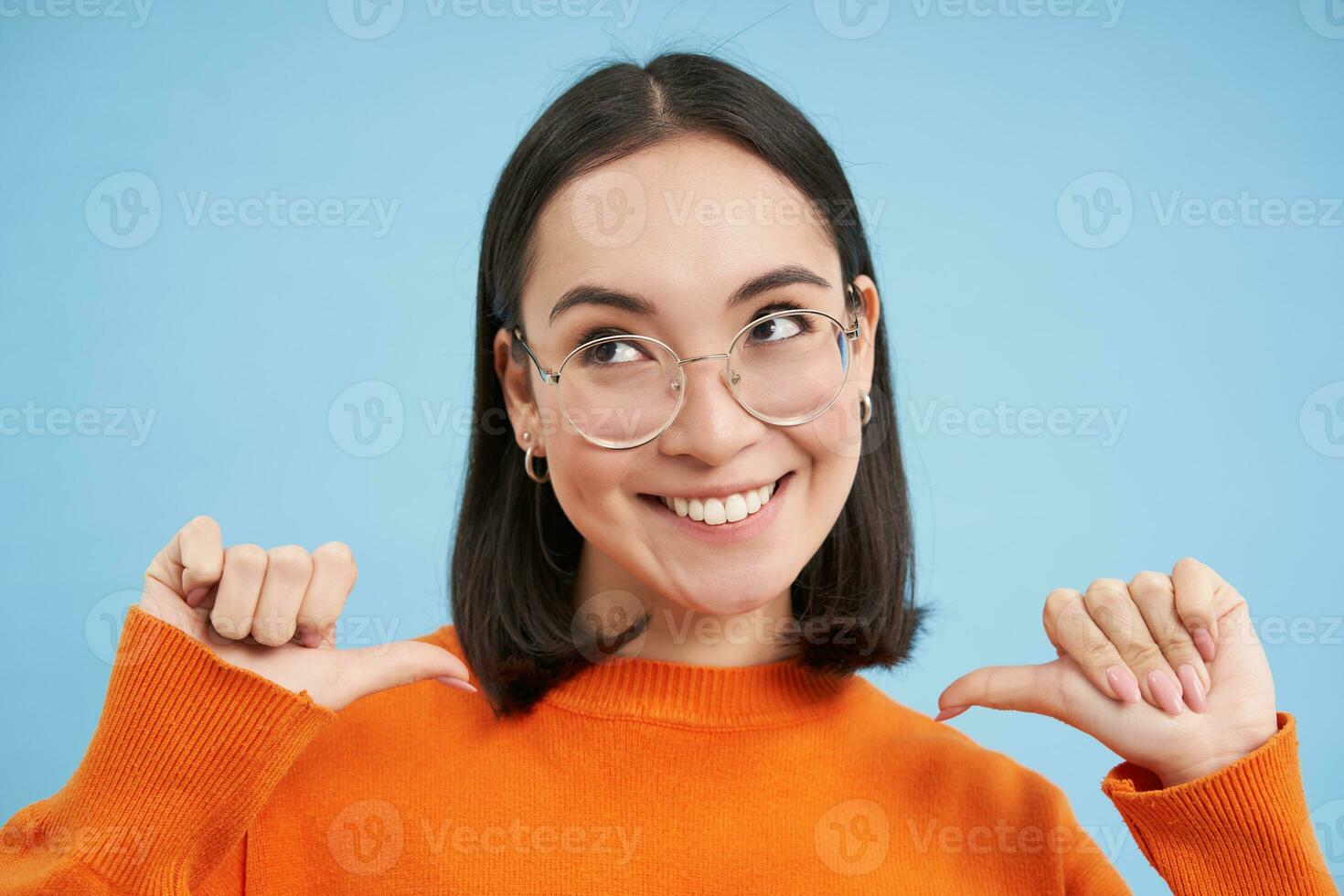 retrato do lindo menina dentro à moda copos, apontando às ela mesma com confiança, ser autoconfiante, em pé sobre azul fundo foto