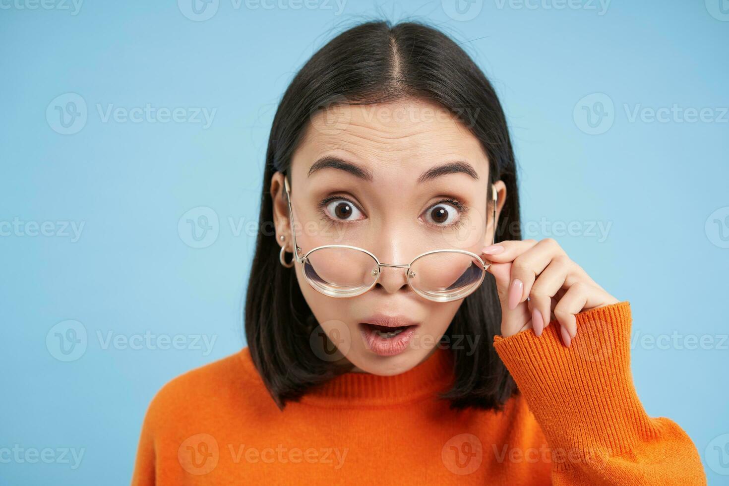retrato do japonês garota, leva do copos, olhares fixos às Câmera dentro óculos, parece espantado, azul fundo foto