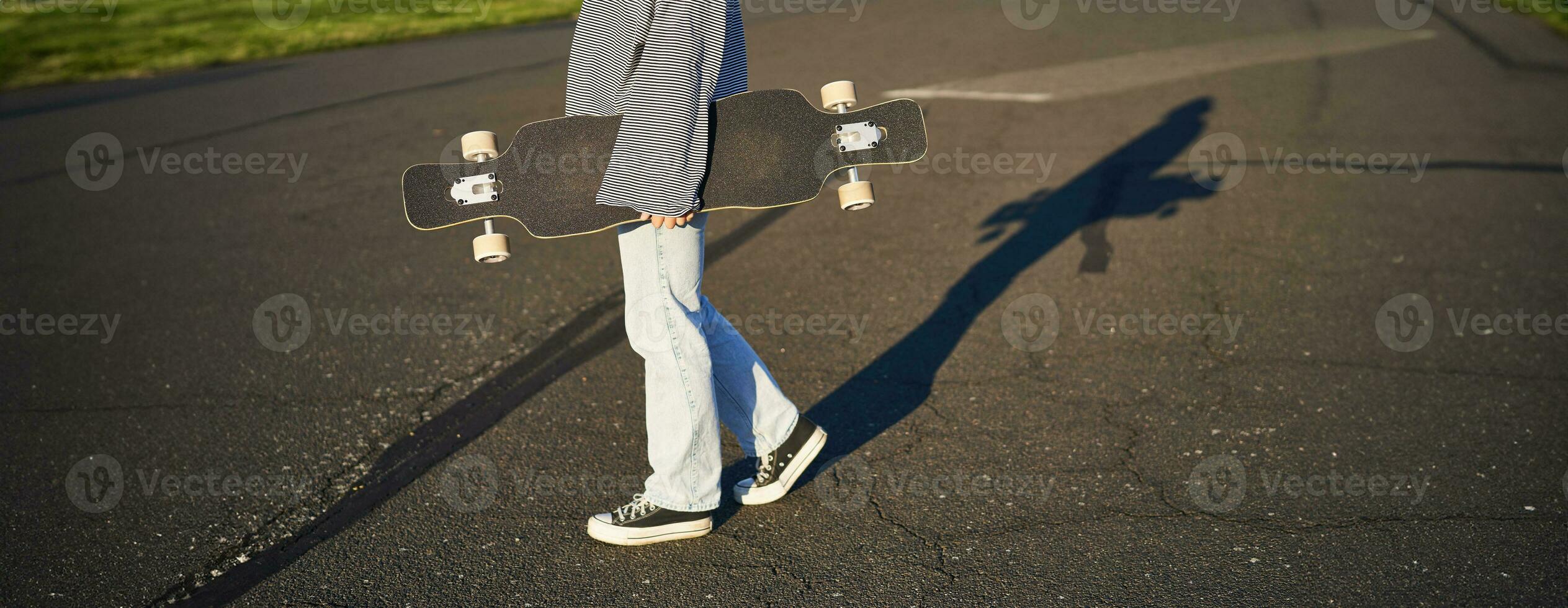 cortada tiro do adolescente menina corpo, segurando cruzador Longboard dentro mão, caminhando dentro tênis em estrada dentro jeans e suéter. jovem mulher skatista com skate foto