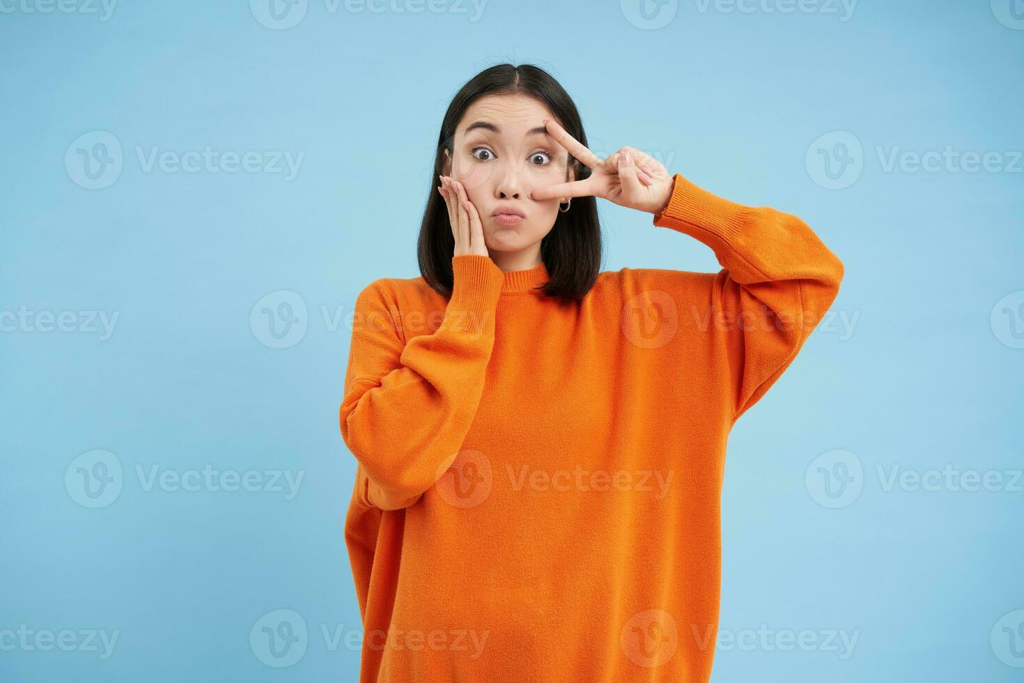 fofa jovem mulher mostra paz, kawaii gesto e parece boba às Câmera, carrinhos dentro laranja suéter contra azul fundo foto