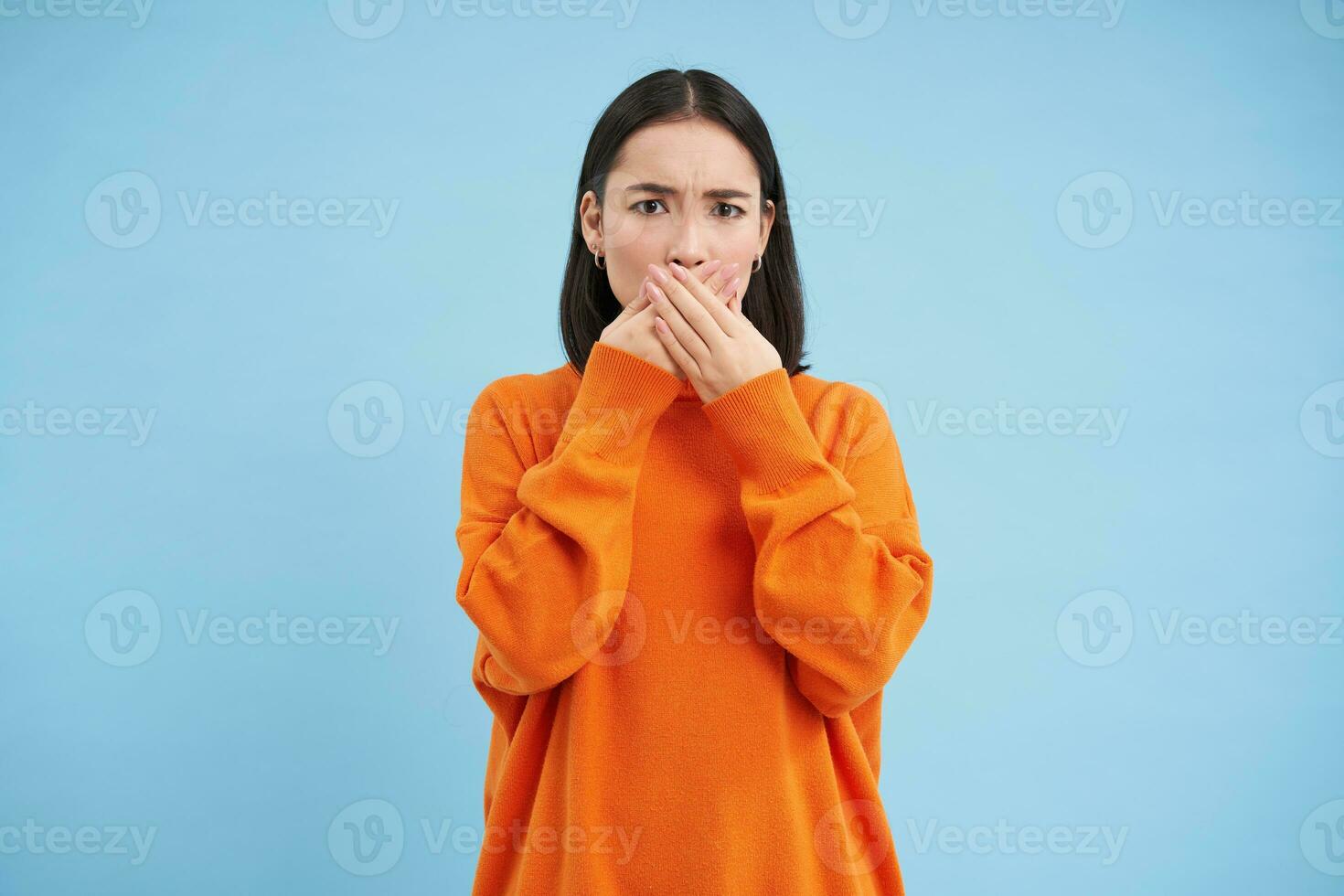 retrato do chocado coreano mulher suspiros, fecha dela boca com mãos e Veja sem palavras às Câmera, carrinhos sobre azul fundo foto