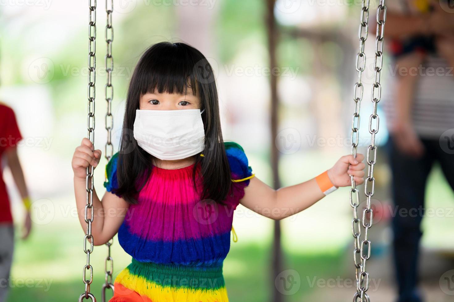linda garota usa máscara médica branca para evitar a propagação do coronavírus covid 19, criança sentada no balanço do playground em um parque lotado, uma criança de 4 anos está usando um vestido colorido, novo normal foto
