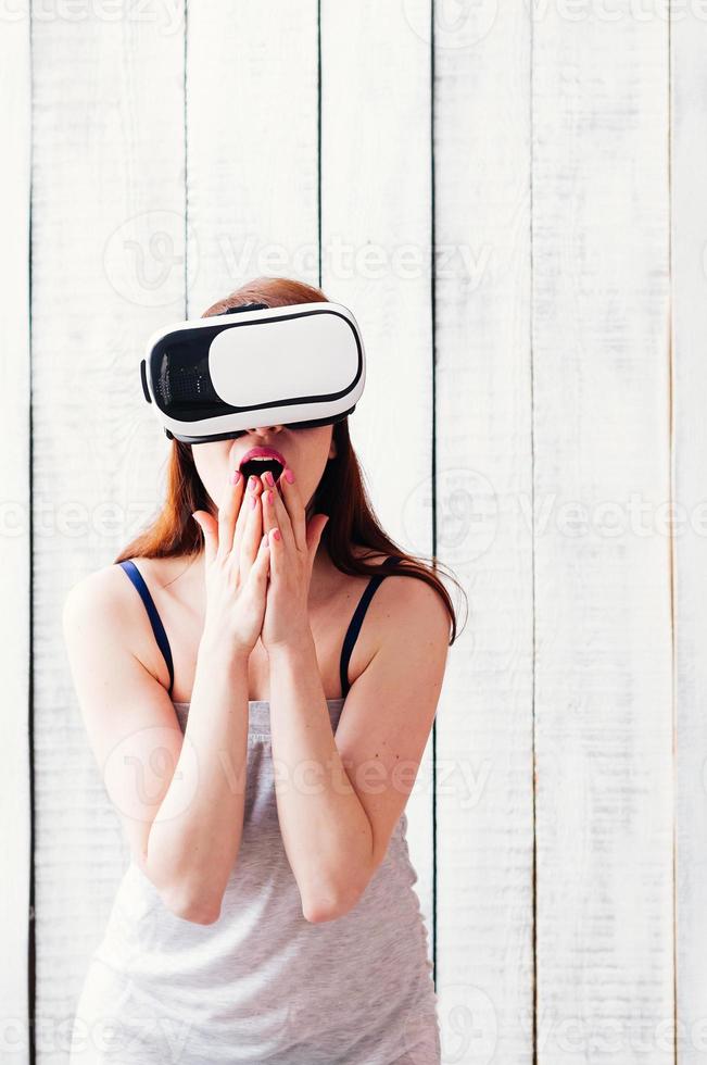 garota de óculos VR, animada, segurando as mãos no rosto, fundo de madeira branco foto