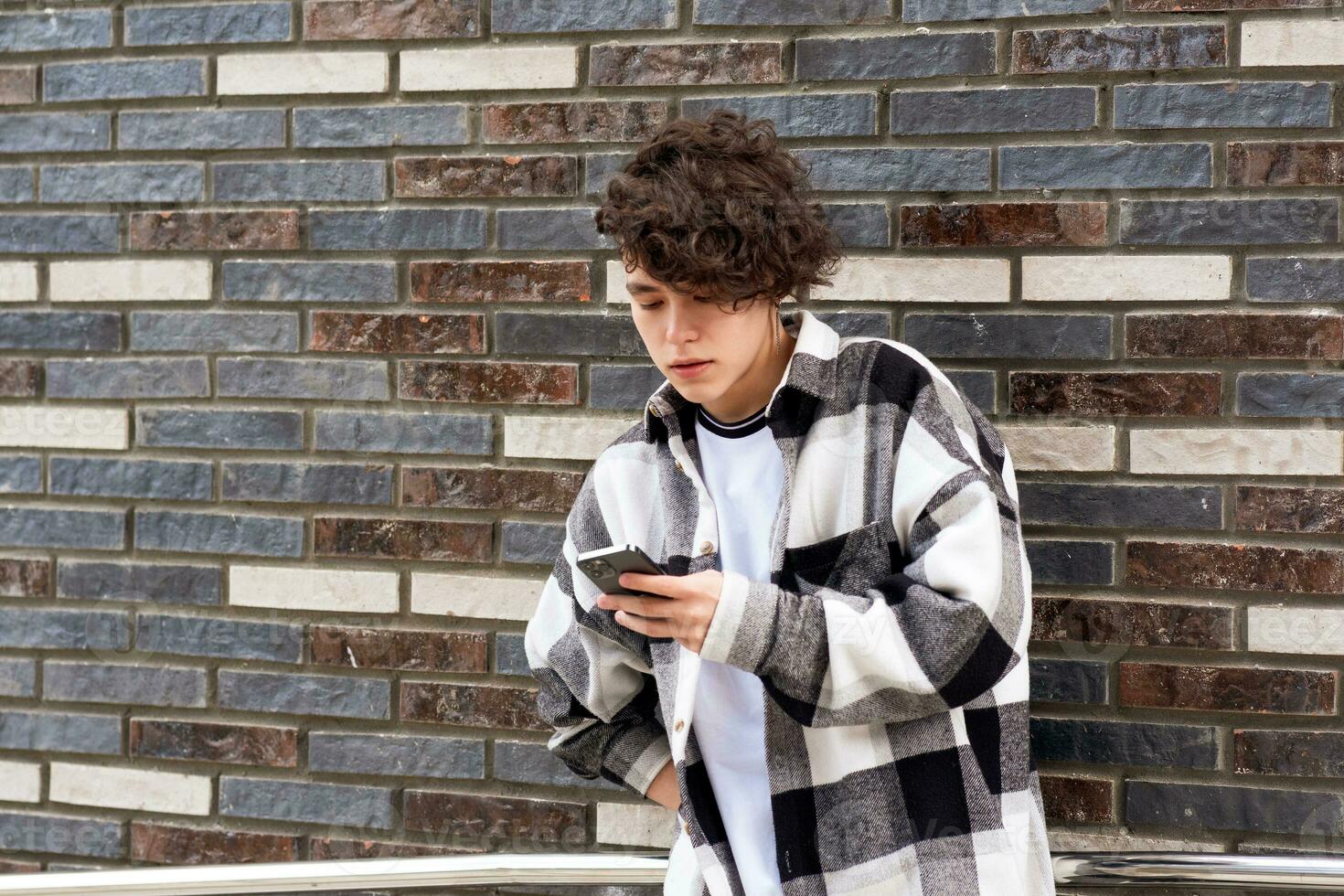 jovem homem lê alguma coisa em a telefone contra uma tijolo parede foto