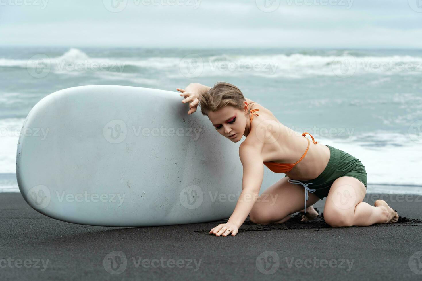 fêmea surfista é ajoelhado em Preto areia do praia, segurando prancha de surfe e relaxado sexy pose foto