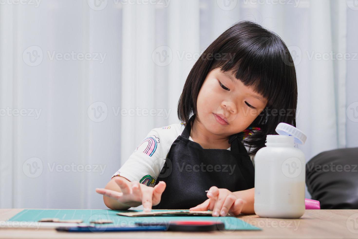 aluno criança garota com papel e cola, fazendo casas de caixa de papelão. criança da escola lição de casa. jovem escolar em casa fazendo artesanato. criança feliz fura a invenção. crianças de 4 a 5 anos. foto