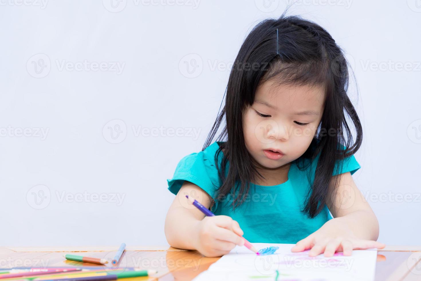 menina asiática está sentada e fazendo lição de casa. criança usa cores de madeira para pintar no livro de desenho. crianças felizes para fazer arte. pratique a concentração e a imaginação. criança tempo independente. bebê de 3-4 anos foto