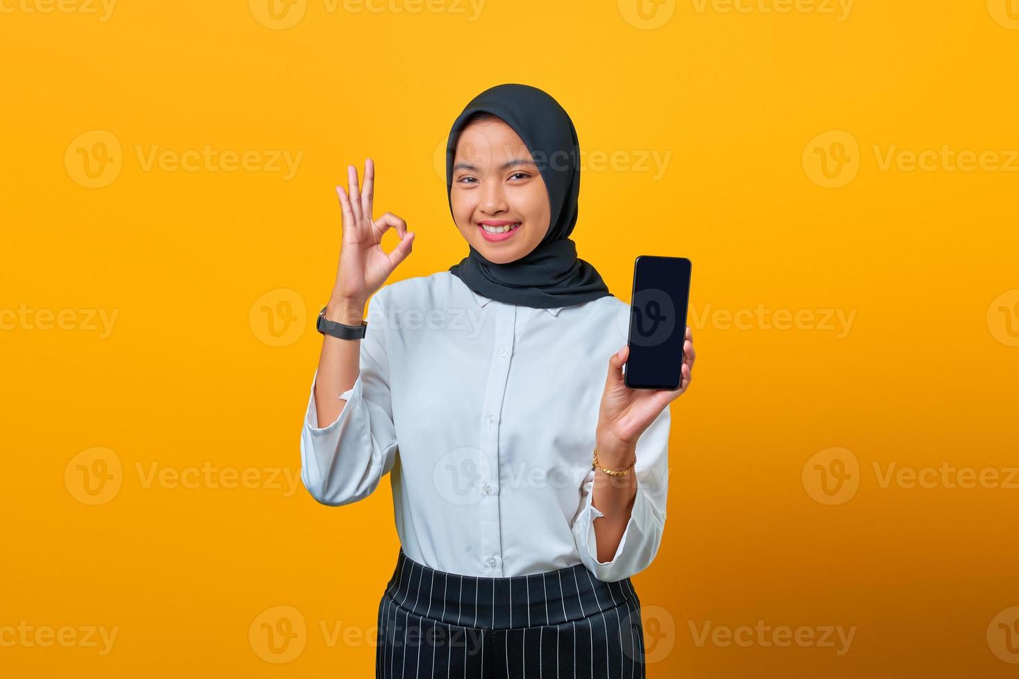Mulher asiática feliz mostrando a tela em branco do celular e um gesto com a mão bem sobre fundo amarelo foto