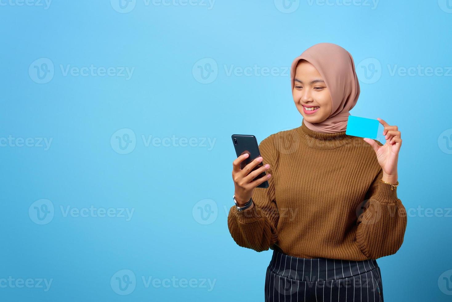jovem mulher asiática feliz mostrando um cartão de crédito e um celular disponível sobre fundo azul foto