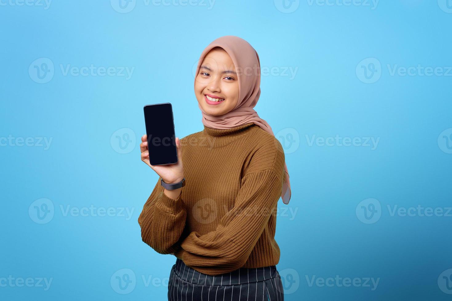 Mulher asiática jovem feliz mostrando a tela em branco do celular isolada sobre um fundo azul foto