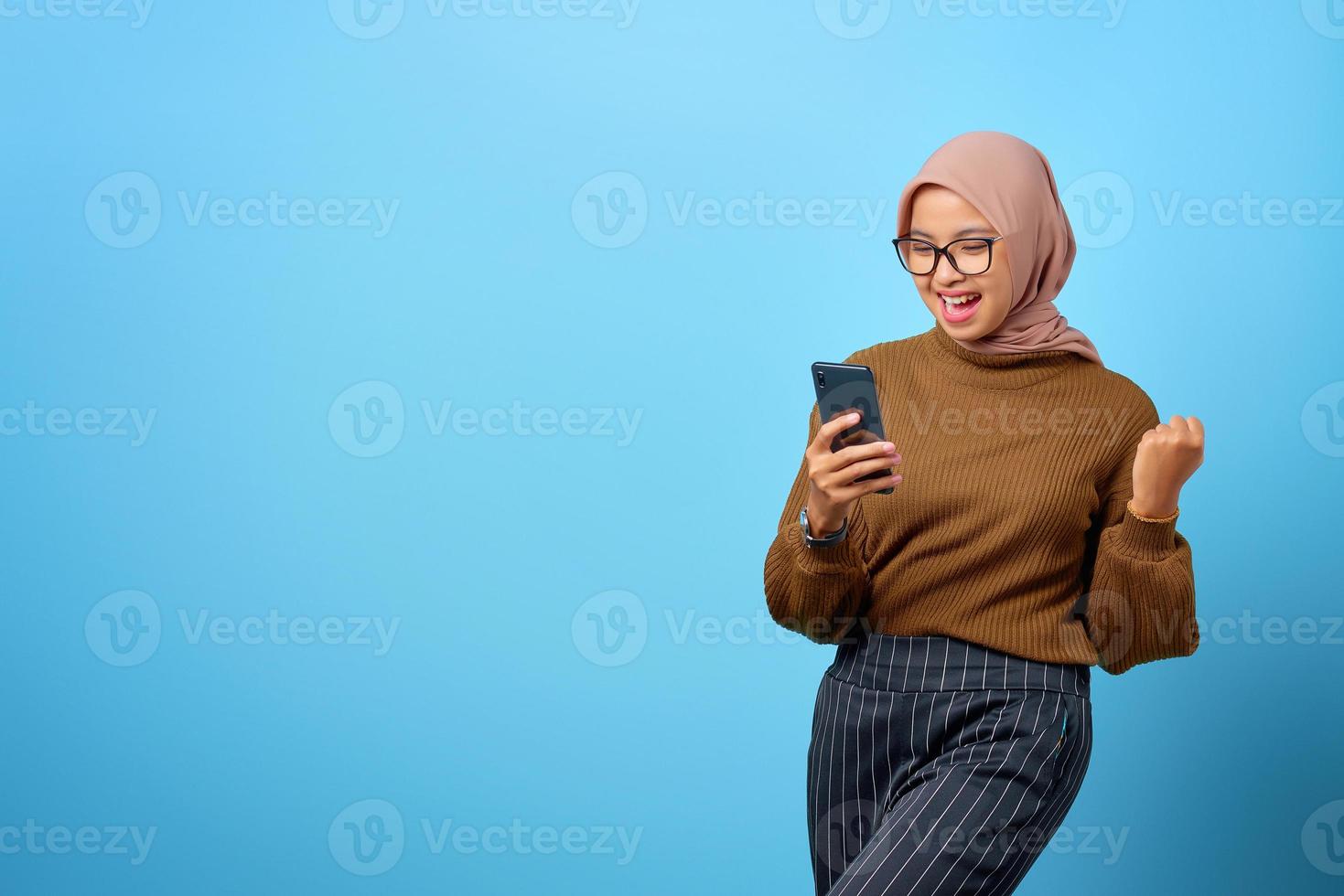 Mulher asiática jovem feliz usando telefone celular com sucesso de gestos com a mão sobre fundo azul foto