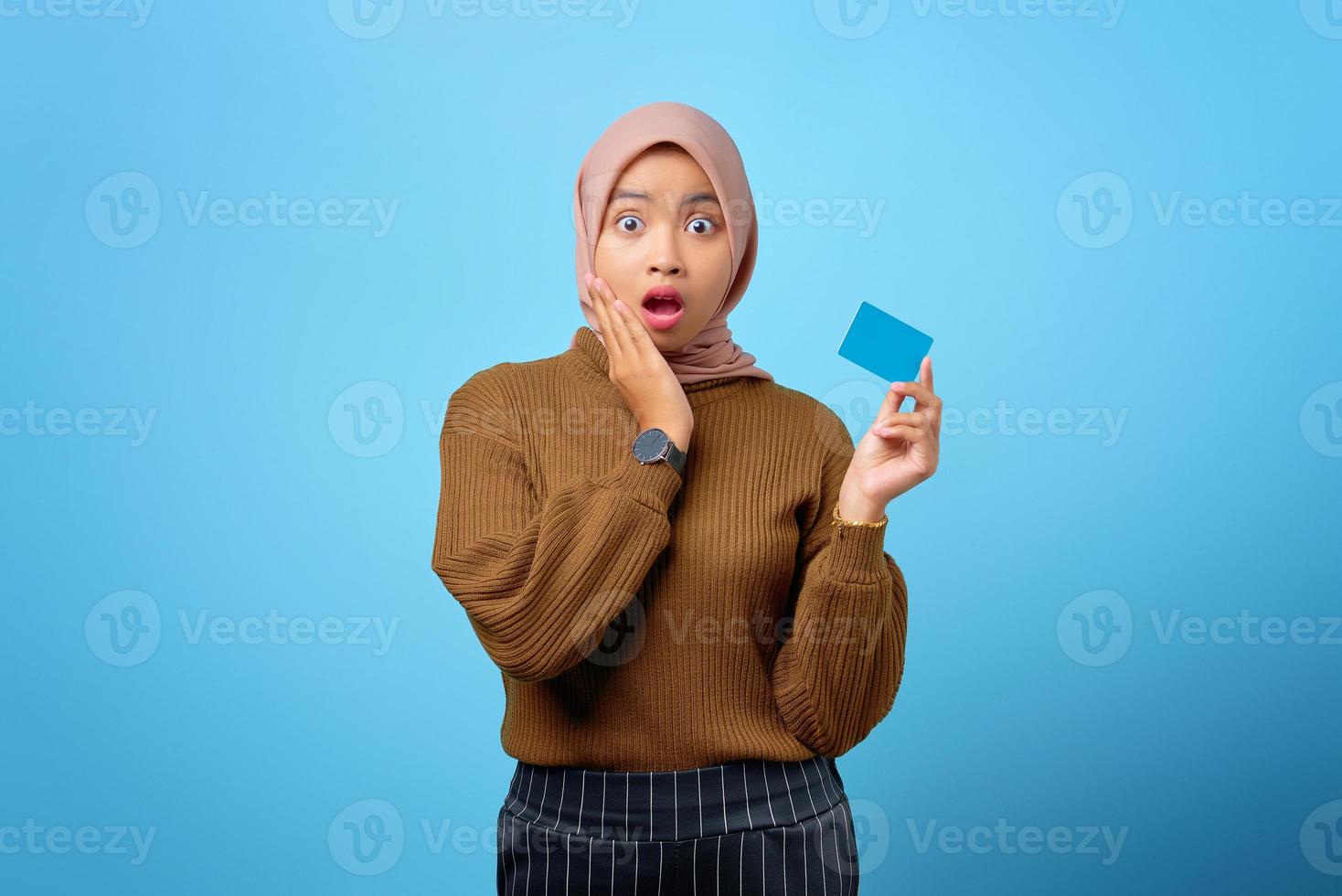 Surpreendeu a jovem mulher asiática segurando um cartão de crédito e tocando as bochechas no fundo azul foto