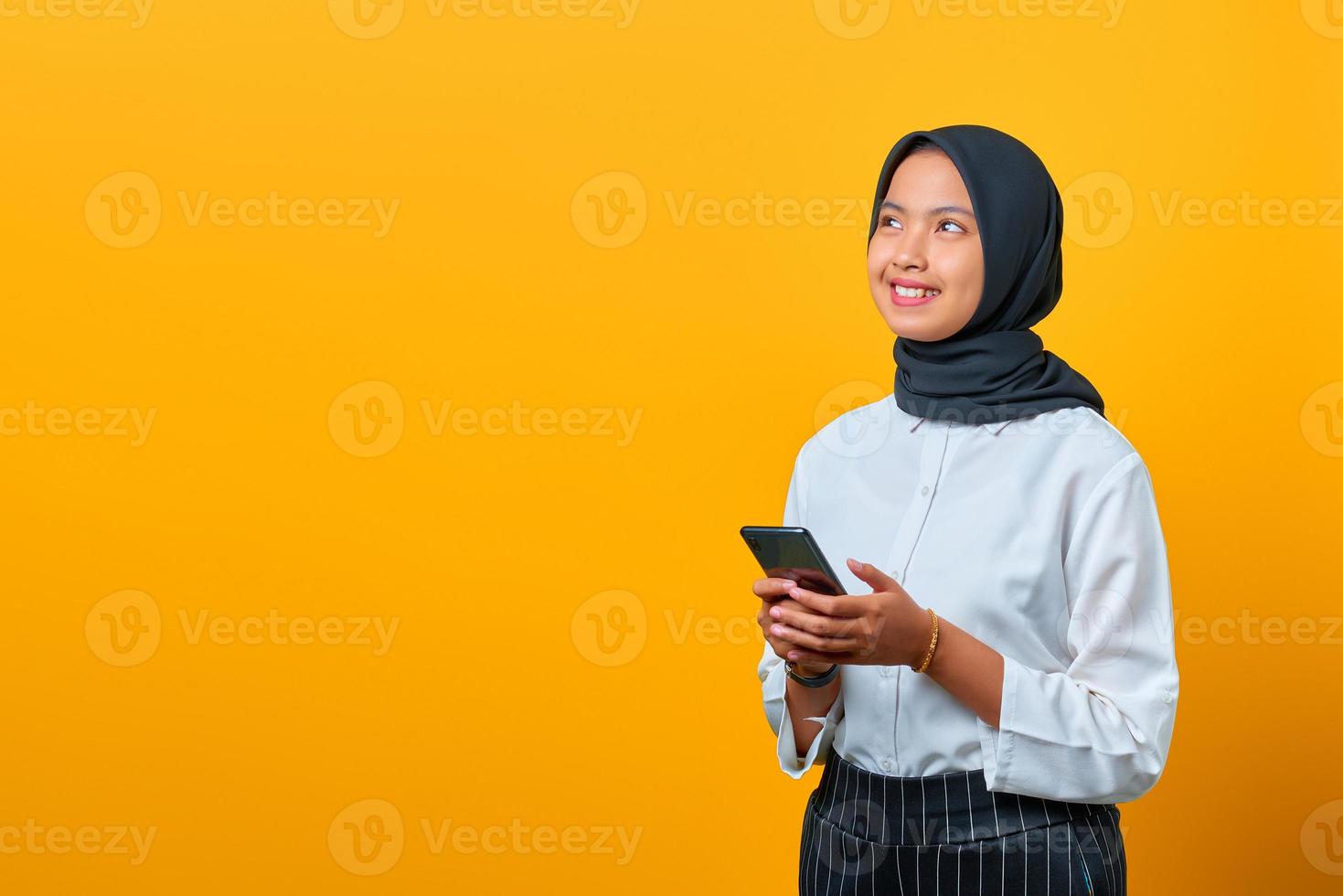 jovem mulher asiática sorridente, usando um telefone celular e olhando de soslaio sobre fundo amarelo foto