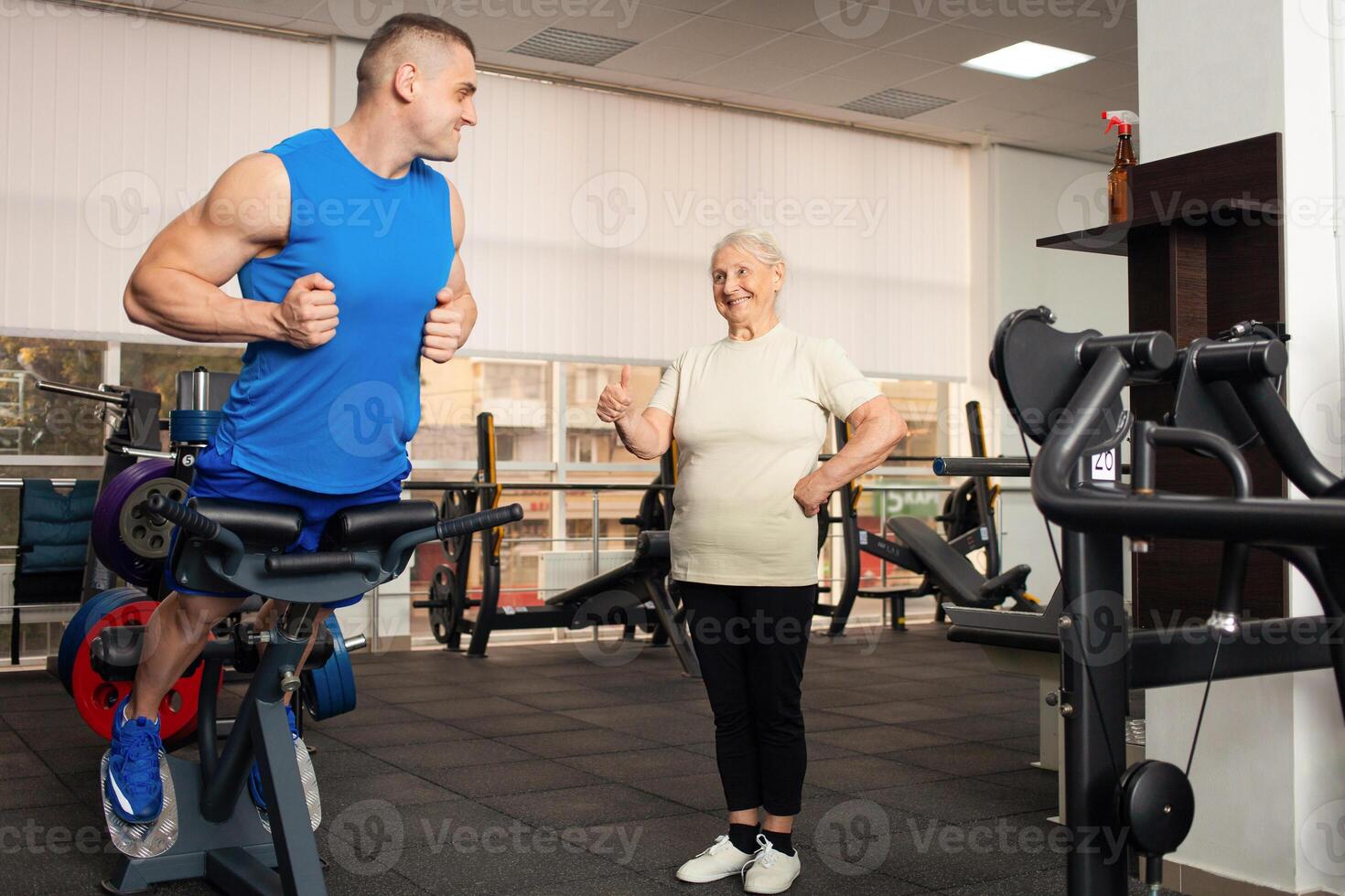 uma lindo jovem treinador mostra a velho mulher quão para Faz a exercício em simulador. pessoas sorriso, estão feliz, mostrar aula, dedão. super. Esportes ativo saudável estilo de vida conceito. pensionista foto