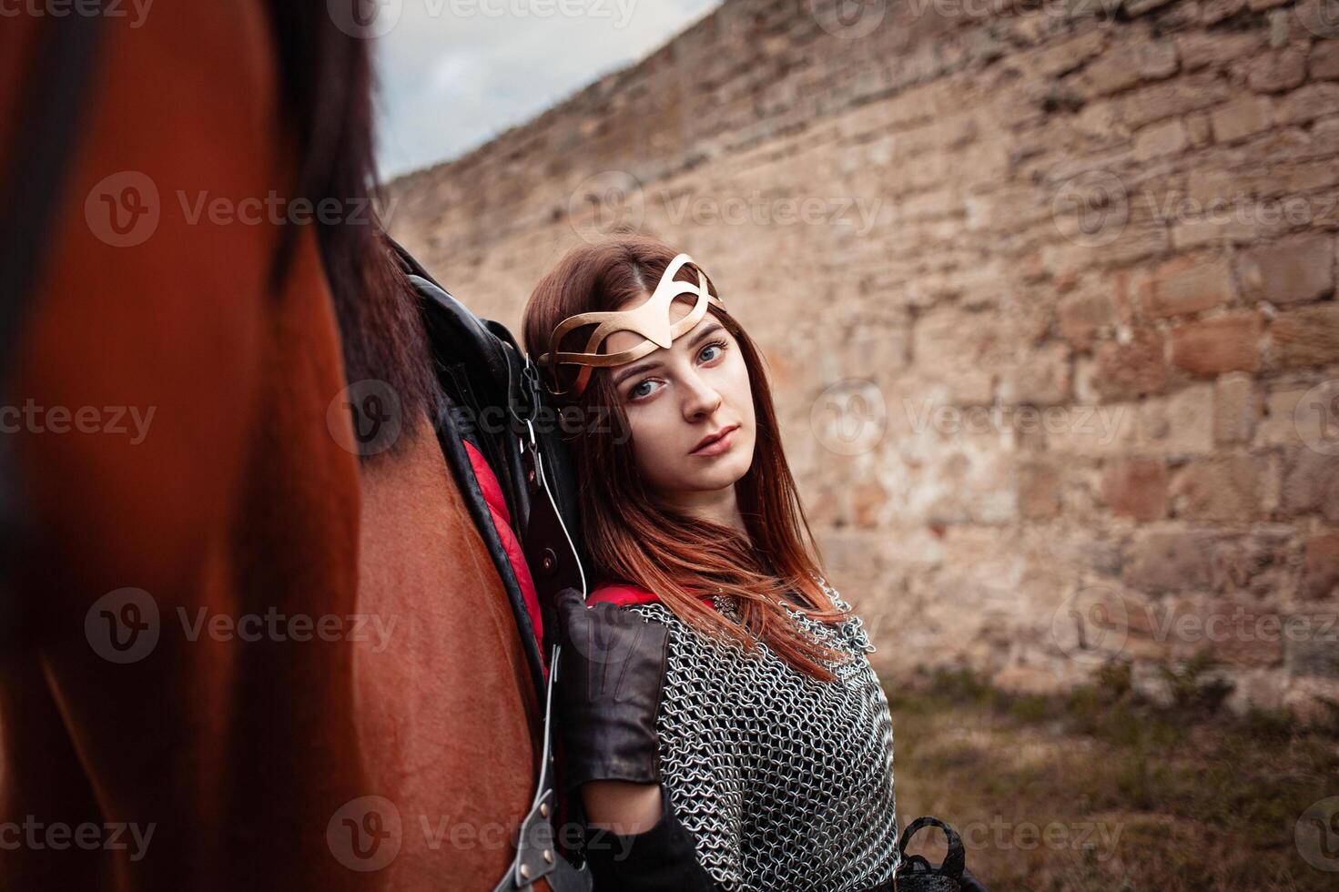 uma lindo menina carrinhos Próximo para uma cavalo contra uma pedra muro. uma mulher vestido Como uma Guerreiro rainha. foto