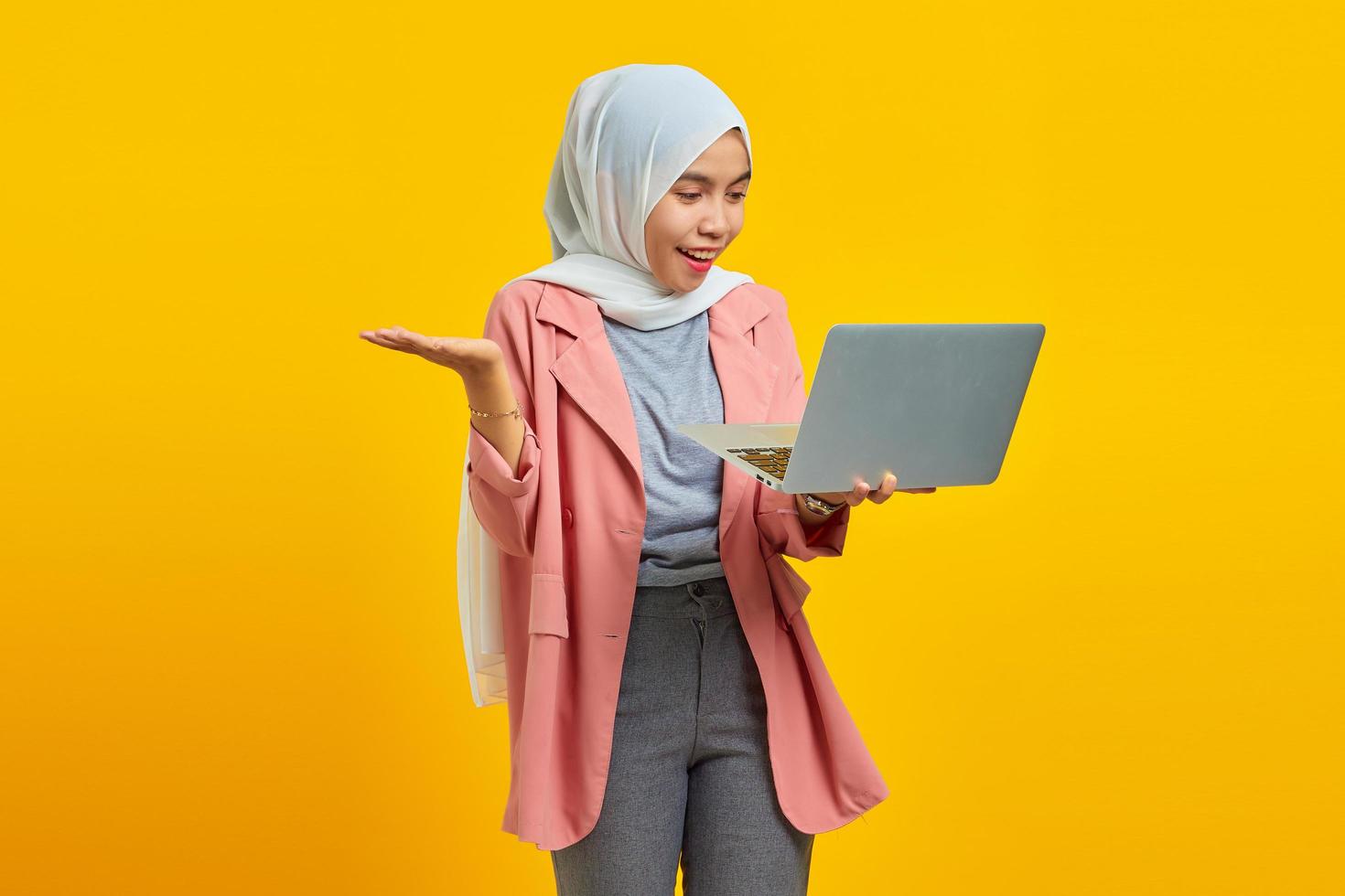 retrato de uma jovem asiática alegre segurando um laptop e apontando isolado sobre um fundo amarelo foto