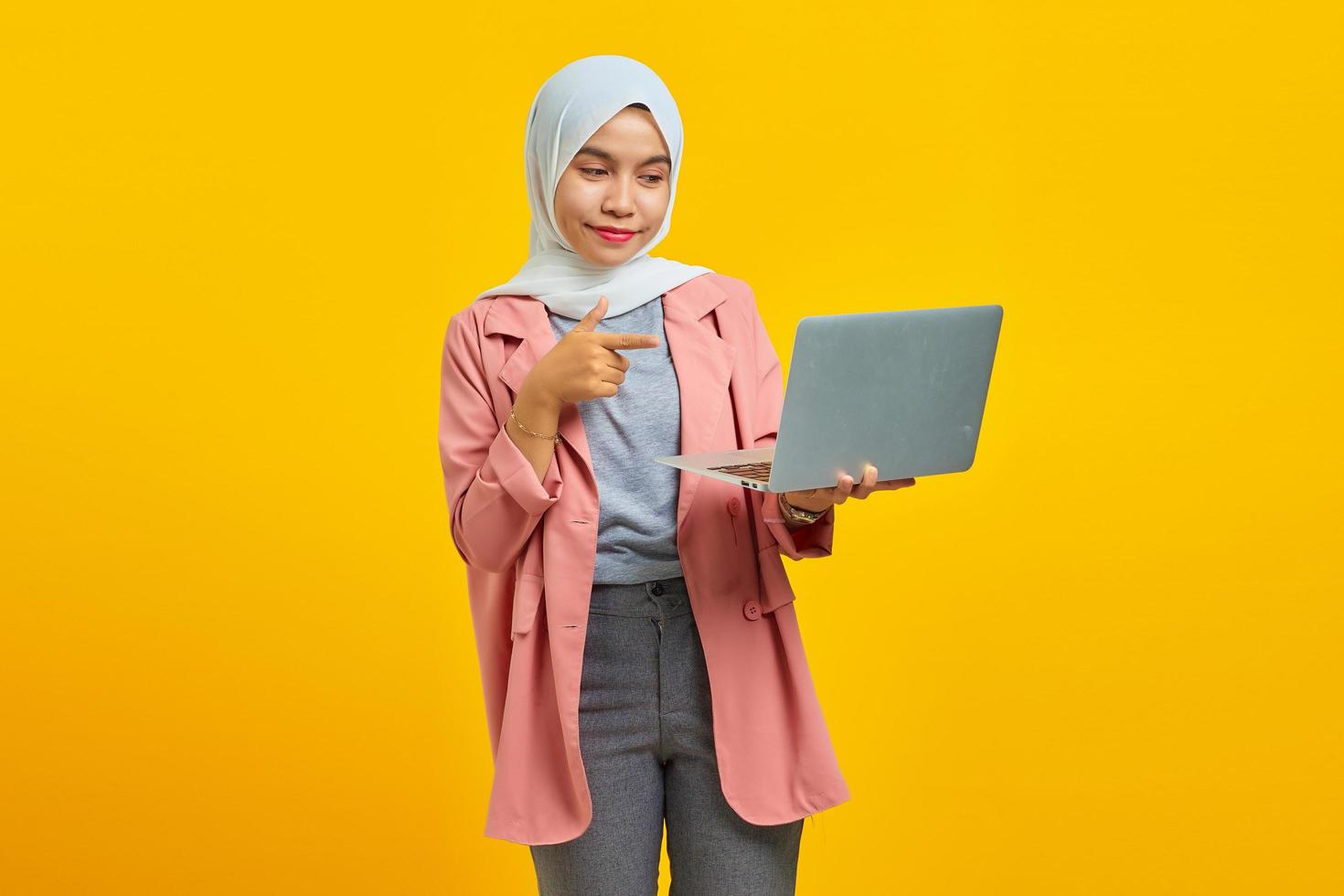 retrato de uma jovem asiática sorridente segurando um laptop e apontando isolado sobre um fundo amarelo foto