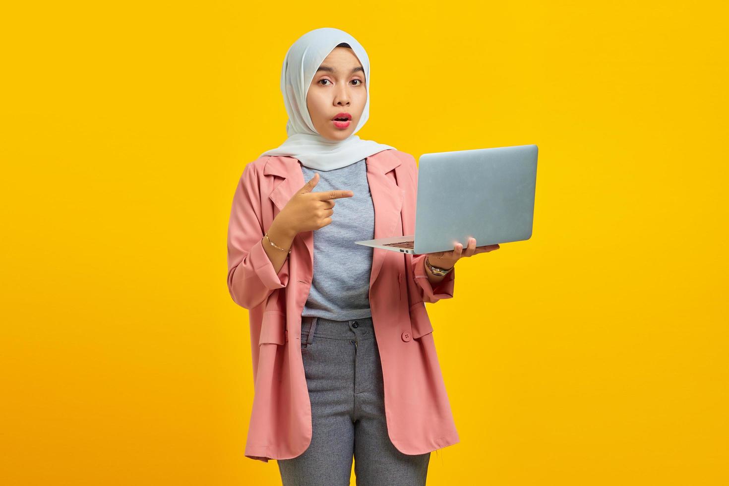 retrato de uma jovem asiática animada segurando um laptop e apontando isolado sobre um fundo amarelo foto