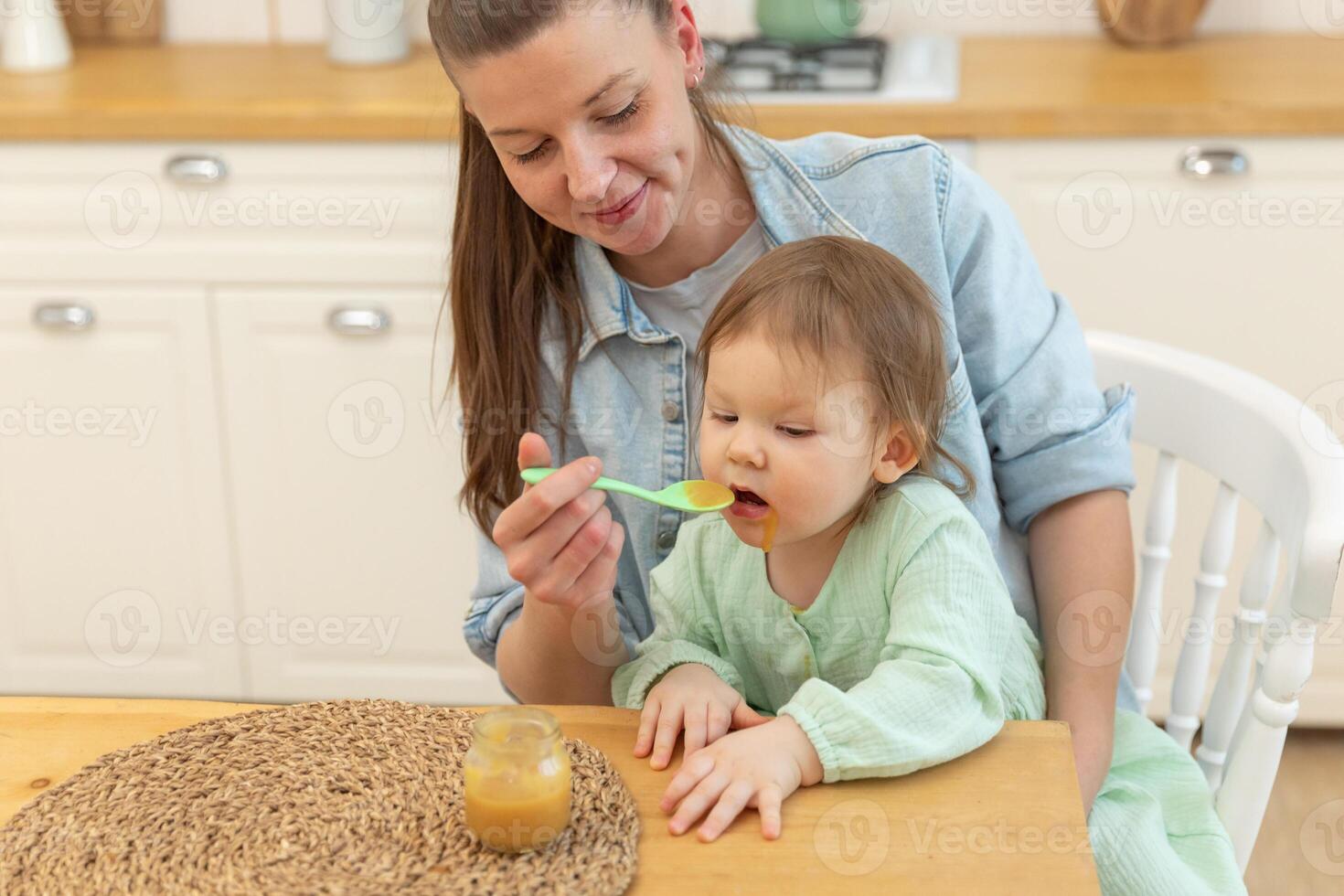 feliz família às lar. mãe alimentando dela bebê menina a partir de colher dentro cozinha. pequeno criança pequena criança com bagunçado engraçado face come saudável Comida às lar. jovem mulher mãe dando Comida para criança filha foto