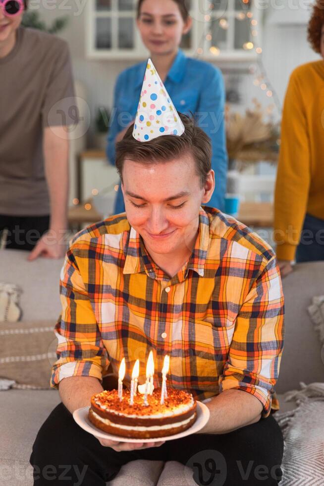 faço uma desejar. homem vestindo festa boné sopro Fora queimando velas em aniversário bolo. feliz aniversário Festa. grupo do amigos desejos cara feliz aniversário. pessoas a comemorar aniversário com festa às casa foto