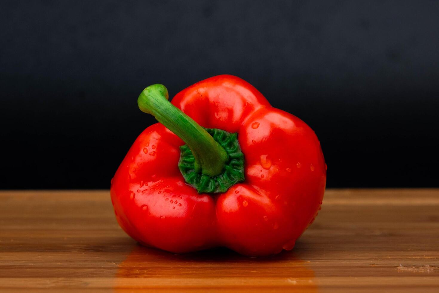 fresco vermelho Sino Pimenta com água gotas uma de madeira corte borda em uma Preto fundo. foto