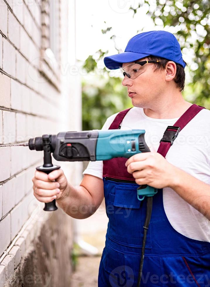 trabalhador da construção civil em óculos de proteção e uniforme com perfurador de perfuração da parede ao ar livre. homem com broca foto