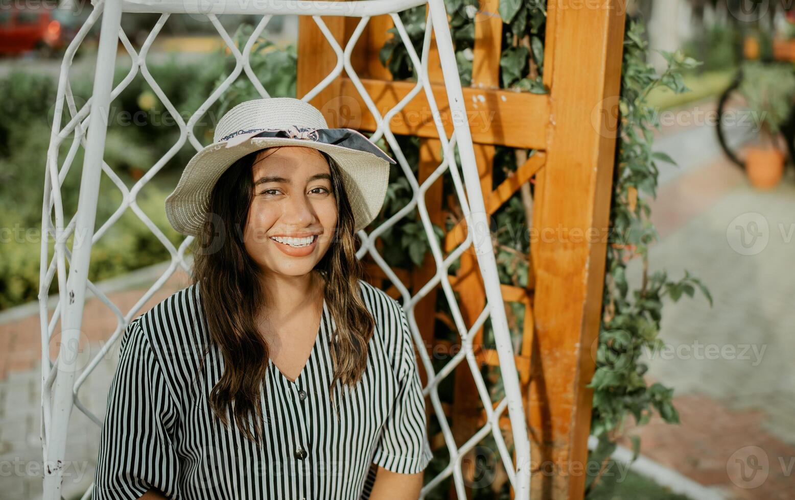 retrato do uma sorridente menina dentro uma chapéu sentado em uma branco balanço dentro uma jardim. estilo de vida do menina dentro chapéu sentado em uma balanço olhando às Câmera foto