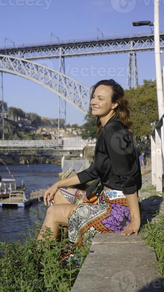 vertical vídeo com lindo mulher em aterro. Ação. atraente mulher é sentado em Beira do aterro em ensolarado verão dia. à moda mulher é em repouso em cidade aterro com Visão do ponte foto