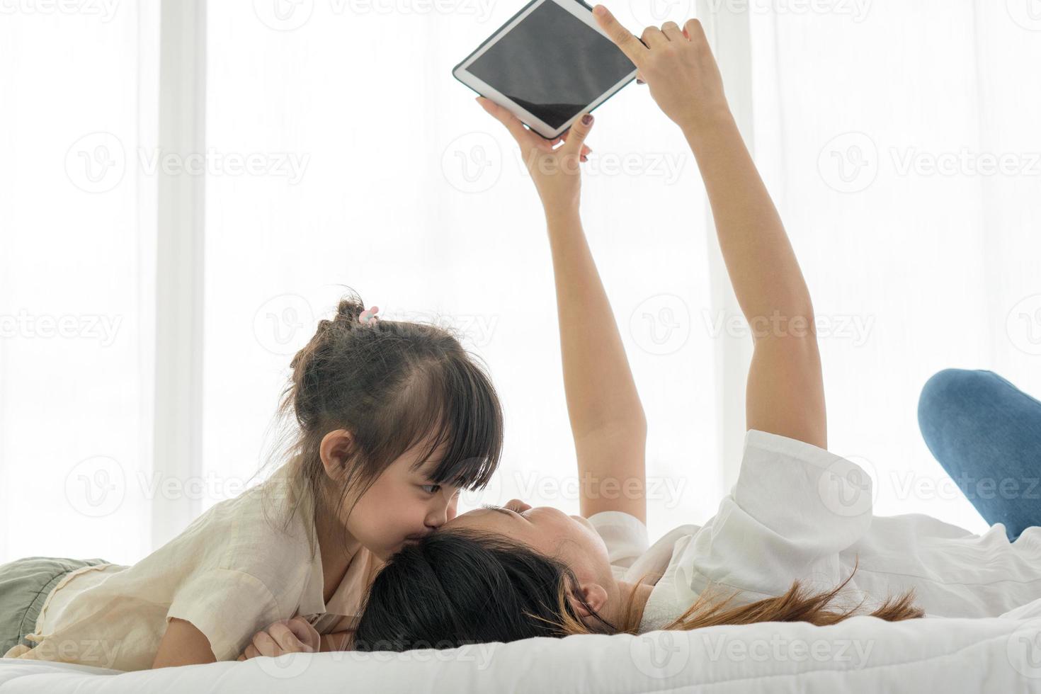 criança asiática beijando a mãe dela enquanto a mãe usando o tablet para tirar foto deles na cama. conceito de família unida