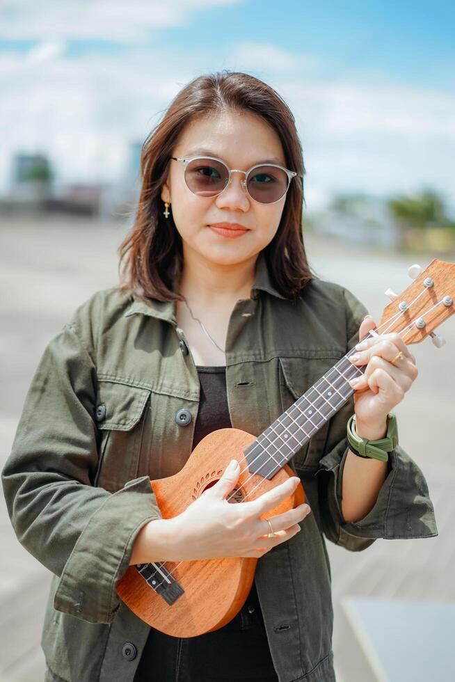 jogando ukulele do jovem lindo ásia mulher vestindo Jaqueta e Preto jeans posando ao ar livre foto