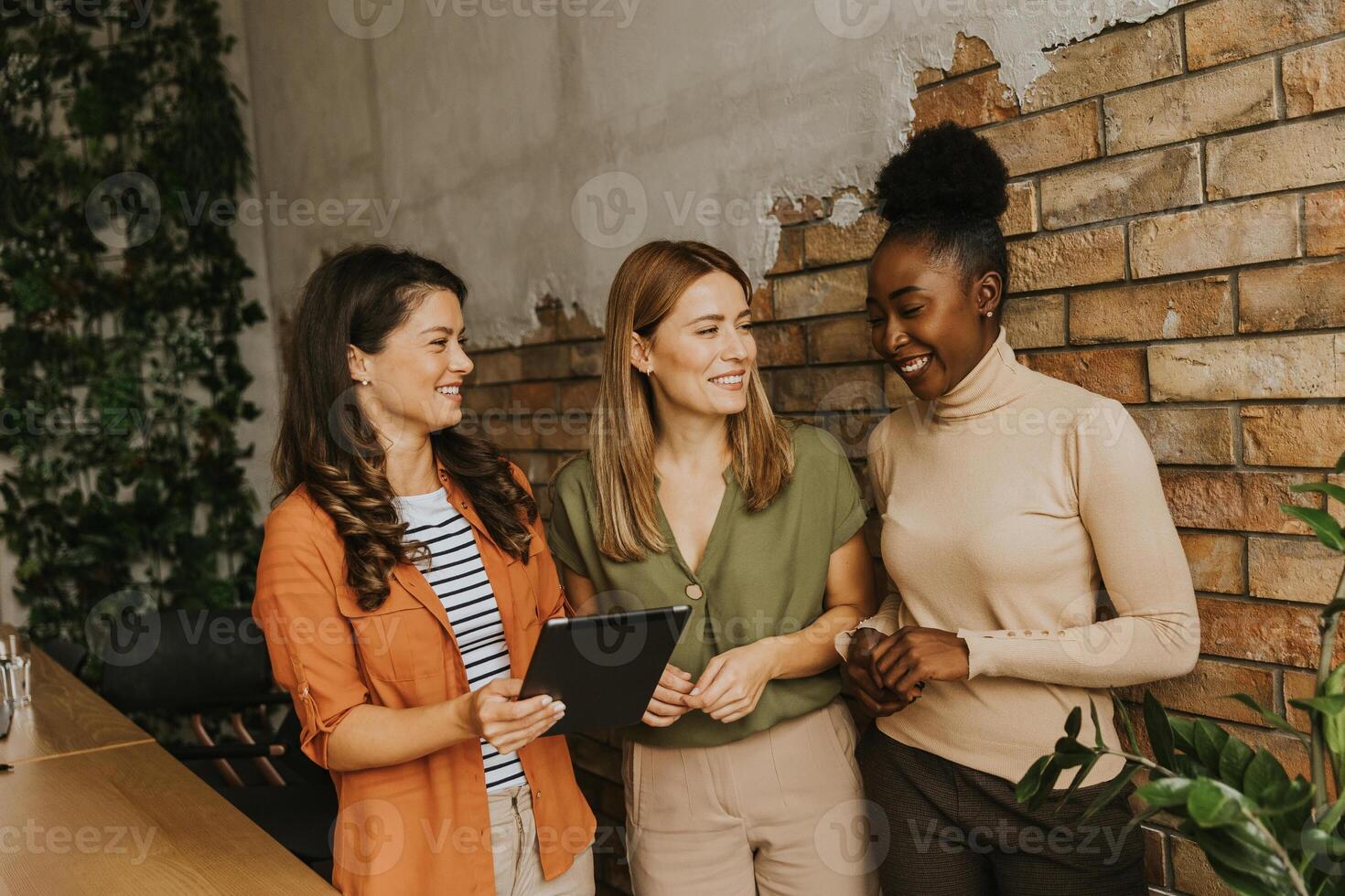 três jovem o negócio mulheres com digital tábua em pé de a tijolo parede dentro a industrial estilo escritório foto