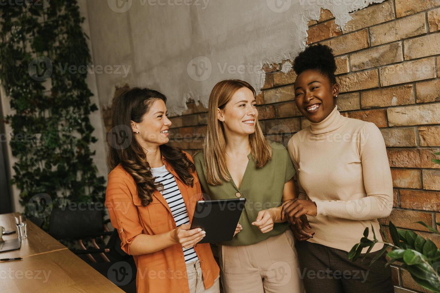 três jovem o negócio mulheres com digital tábua em pé de a tijolo parede dentro a industrial estilo escritório foto