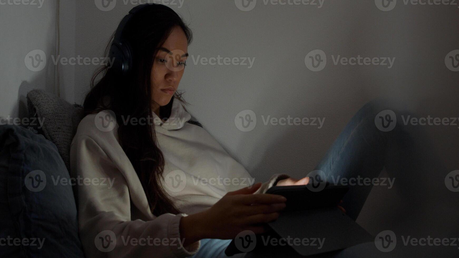 jovem mestiça sentada no sofá no canto da sala, vendo a tela do tablet à sua frente, o reflexo da tela no rosto e nos olhos foto