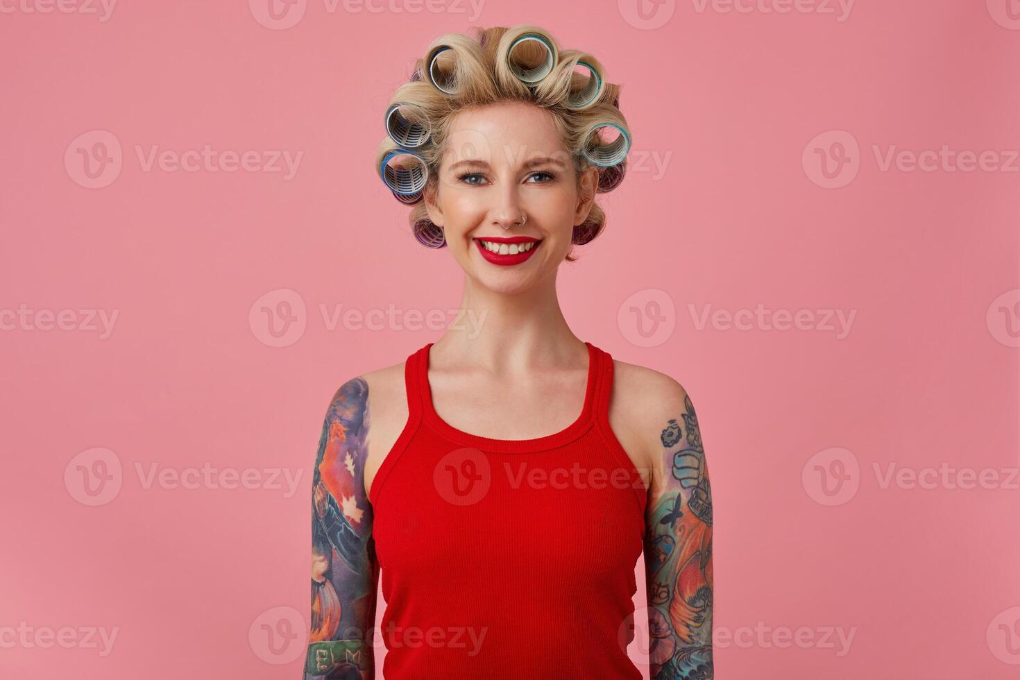 alegre jovem Loiras tatuado fêmea com tarde Maquiagem fazer penteado enquanto posando sobre Rosa fundo com mãos abaixo, olhando às Câmera com encantador sorrir foto