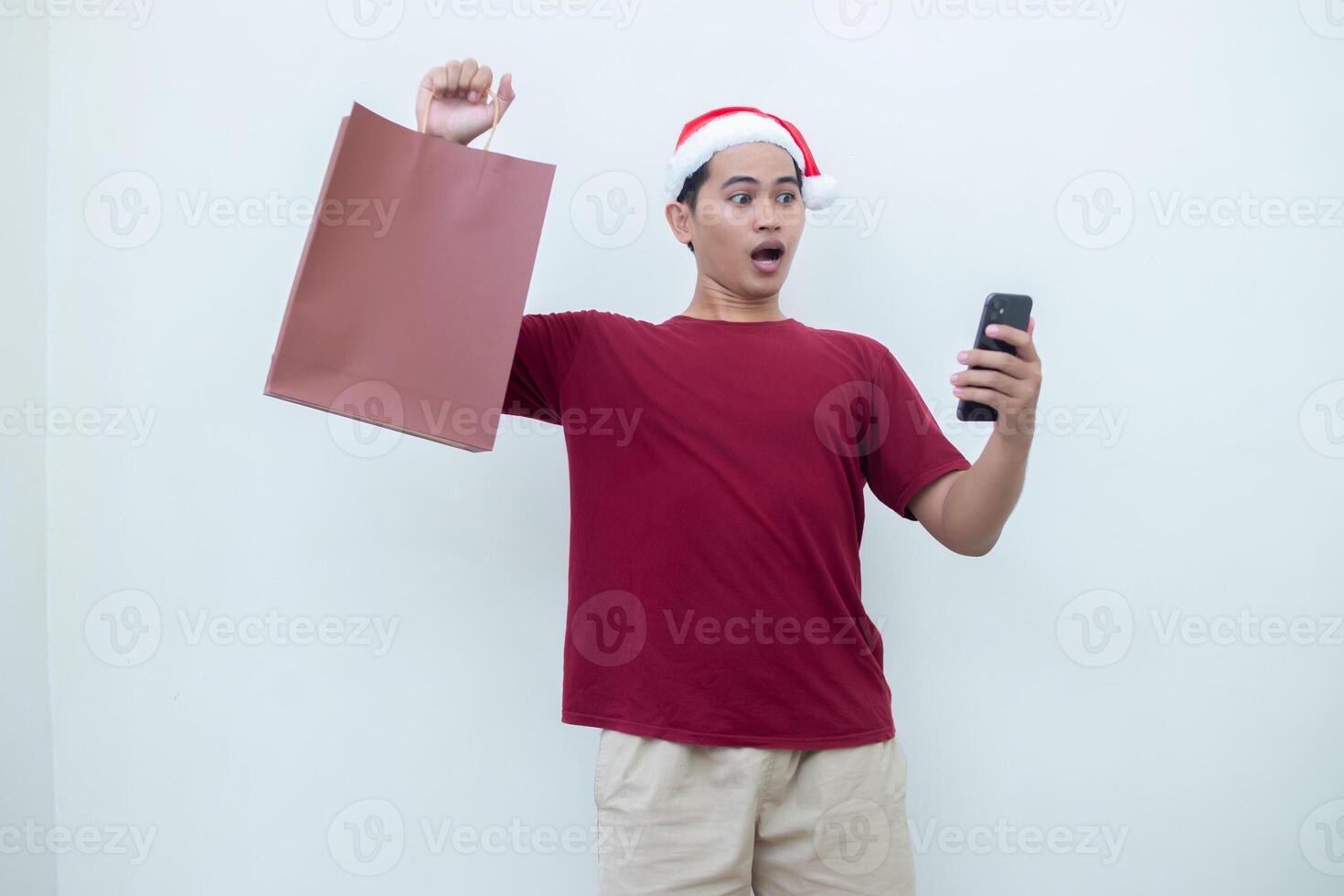 jovem ásia homem vestindo uma santa claus chapéu segurando uma Smartphone e uma compras saco com expressões do sorriso, choque, e surpresa, isolado contra uma branco fundo para visual comunicação foto