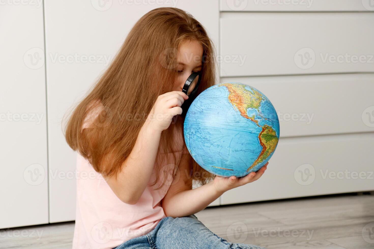 menina sentado em a chão cuidadosamente estudando a globo com uma ampliação vidro foto