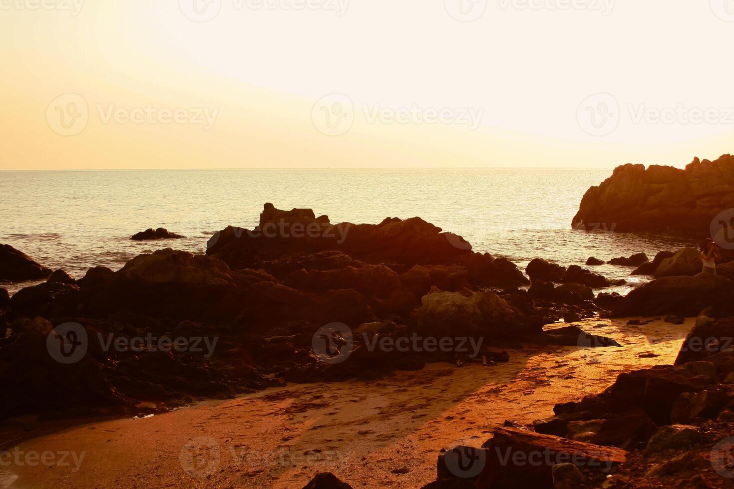 a Sol é configuração em a mar com pedras dentro a primeiro plano, Preto silhuetas em a rochas, luz refletindo fora a mar. foto