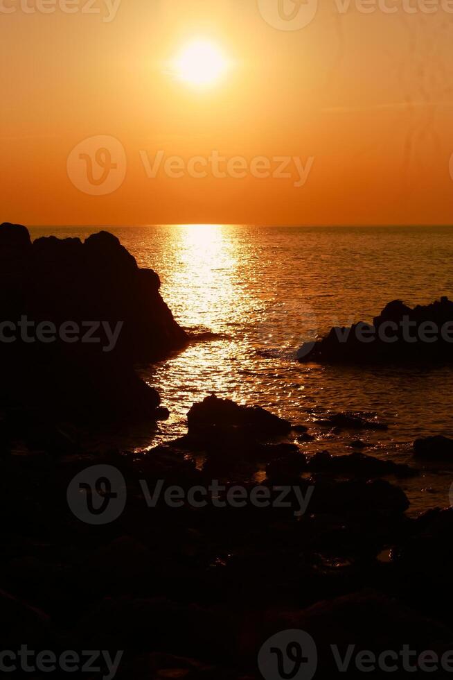 a Sol é configuração em a mar com pedras dentro a primeiro plano, Preto silhuetas em a rochas, luz refletindo fora a mar. foto