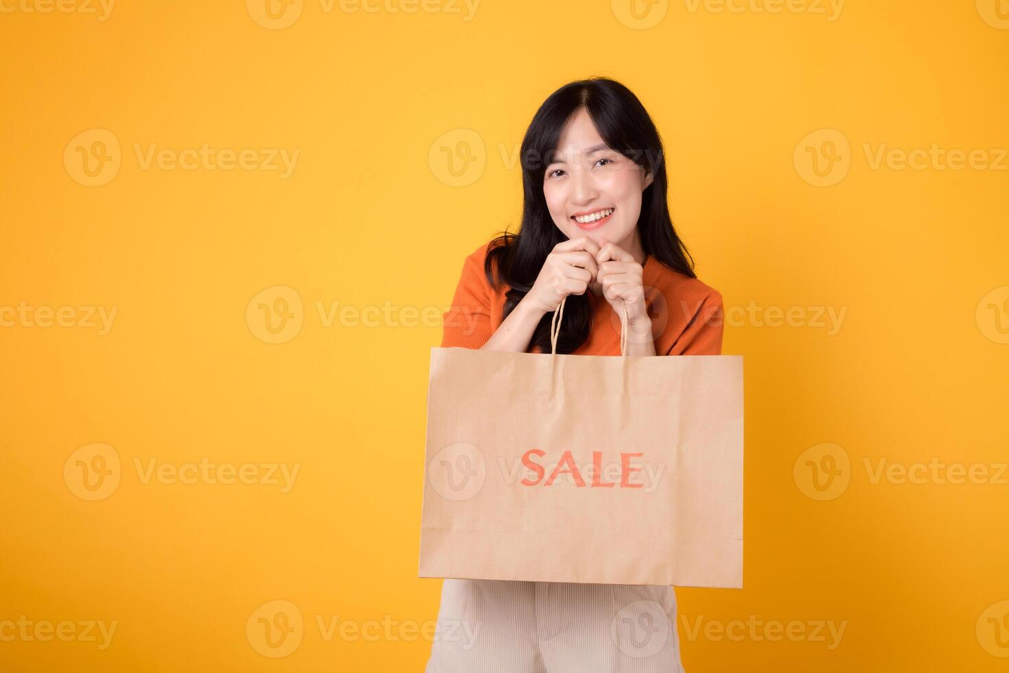 alegre ásia mulher 30s vestindo laranja camisa mostrando feliz sorrir enquanto orgulhosamente exibindo venda papel saco contra amarelo fundo. compras e desfrutando ótimo ofertas conceito. foto