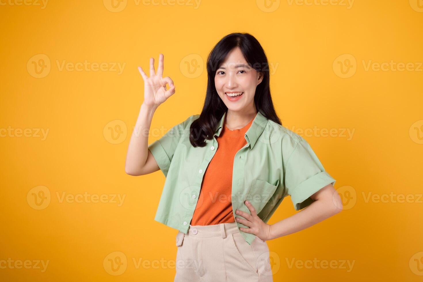 jovem ásia mulher dentro dela 30s, vestido dentro laranja camisa e verde saltador. dela OK mão gesto e □ Gentil sorriso, isolado em uma amarelo fundo, transmitir uma positivo mensagem através corpo linguagem. foto