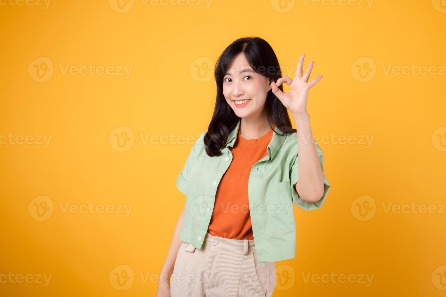 encantador jovem ásia mulher 30s, elegantemente vestido dentro laranja camisa e verde saltador. dela cativante OK mão gesto e □ Gentil sorrir brilho contra uma amarelo fundo, corpo língua conceito. foto