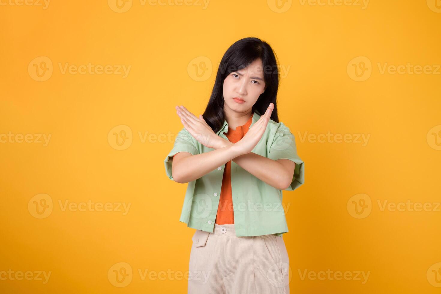 negação com uma cativante jovem ásia mulher 30s, vestindo a laranja camisa e verde saltador. dela Cruz mão gesto, isolado em uma amarelo fundo, representa a conceito do recusa e rejeição. foto