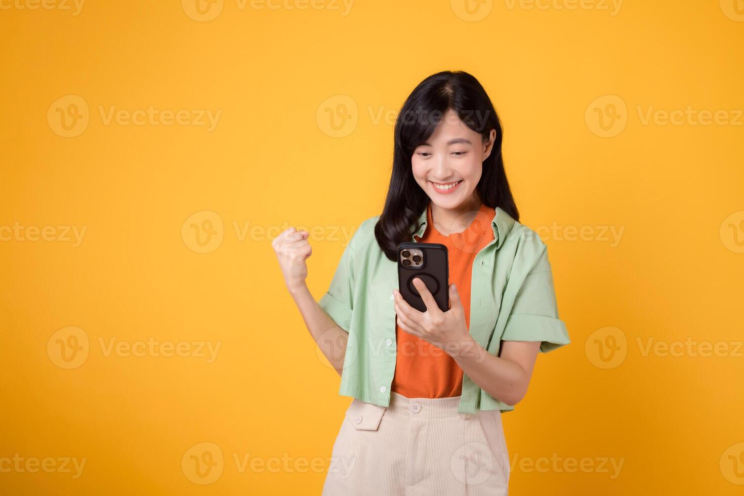 ótimo notícia com uma alegre jovem ásia mulher dentro dela 30s, elegantemente vestido dentro laranja camisa e verde saltador, levantando uma punho com Smartphone em amarelo estúdio fundo. Móvel conceito foto