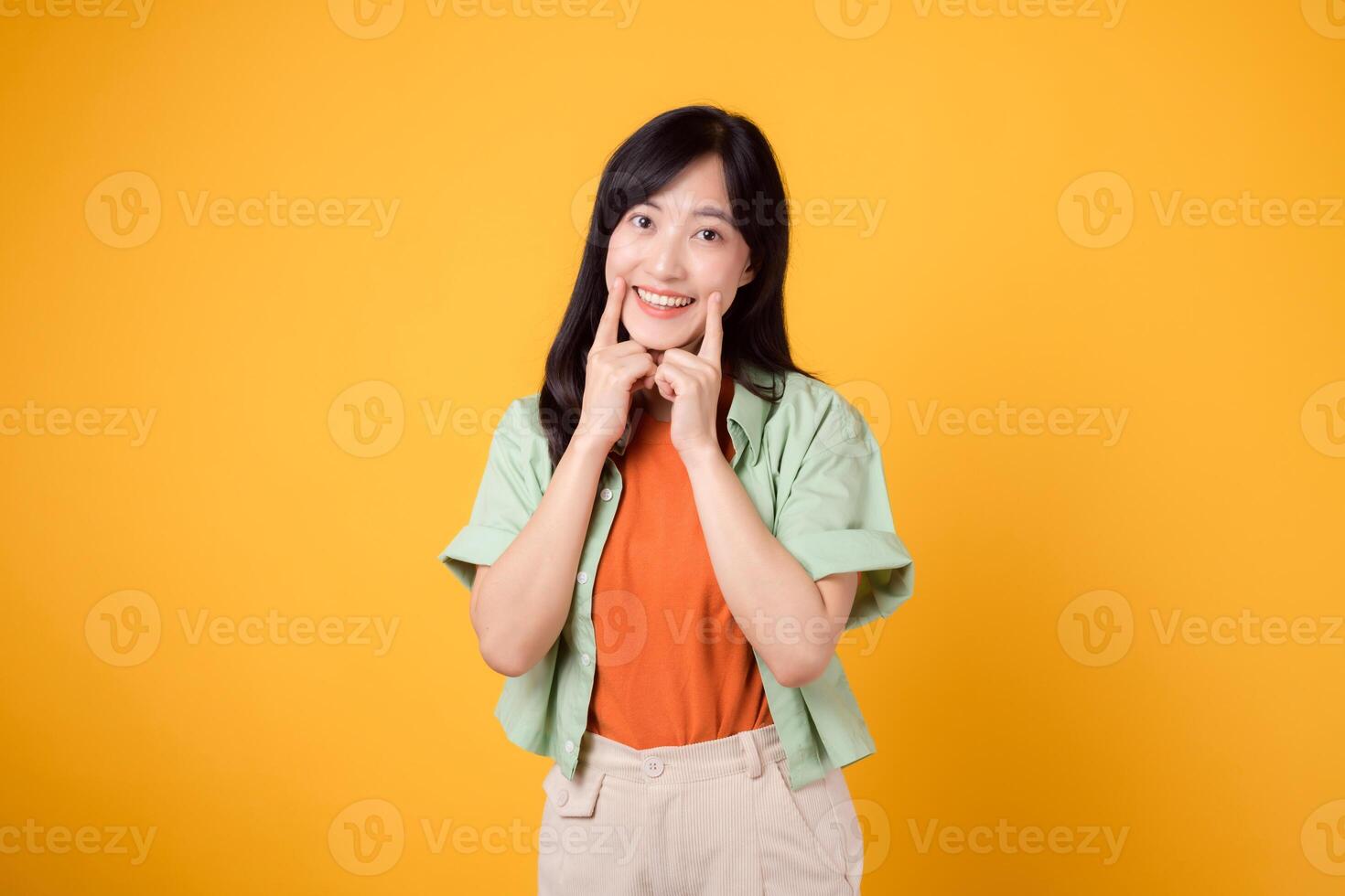 dental bem estar com alegre jovem ásia mulher 30s, elegantemente vestido dentro laranja camisa e verde saltador pontos para dela dentes em amarelo fundo, enfatizando a significado do dental assistência médica. foto