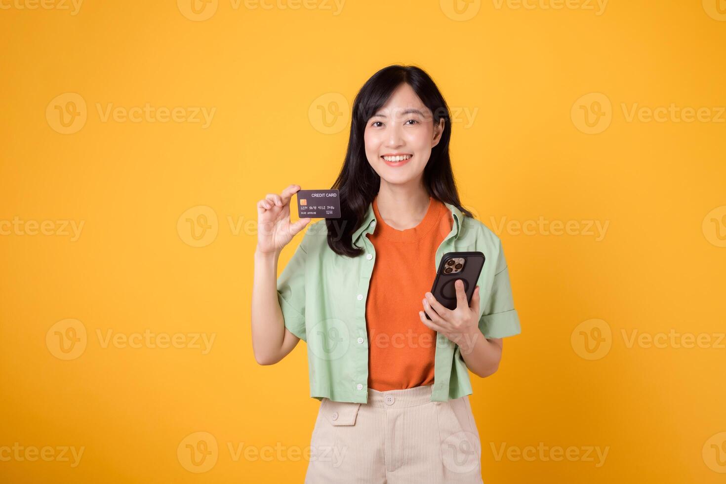 emoção do compras a partir de Móvel com uma cativante jovem ásia mulher dentro dela 30s, elegantemente vestido dentro laranja camisa e verde saltador, usando Smartphone para mostrar crédito cartão em amarelo estúdio fundo. foto