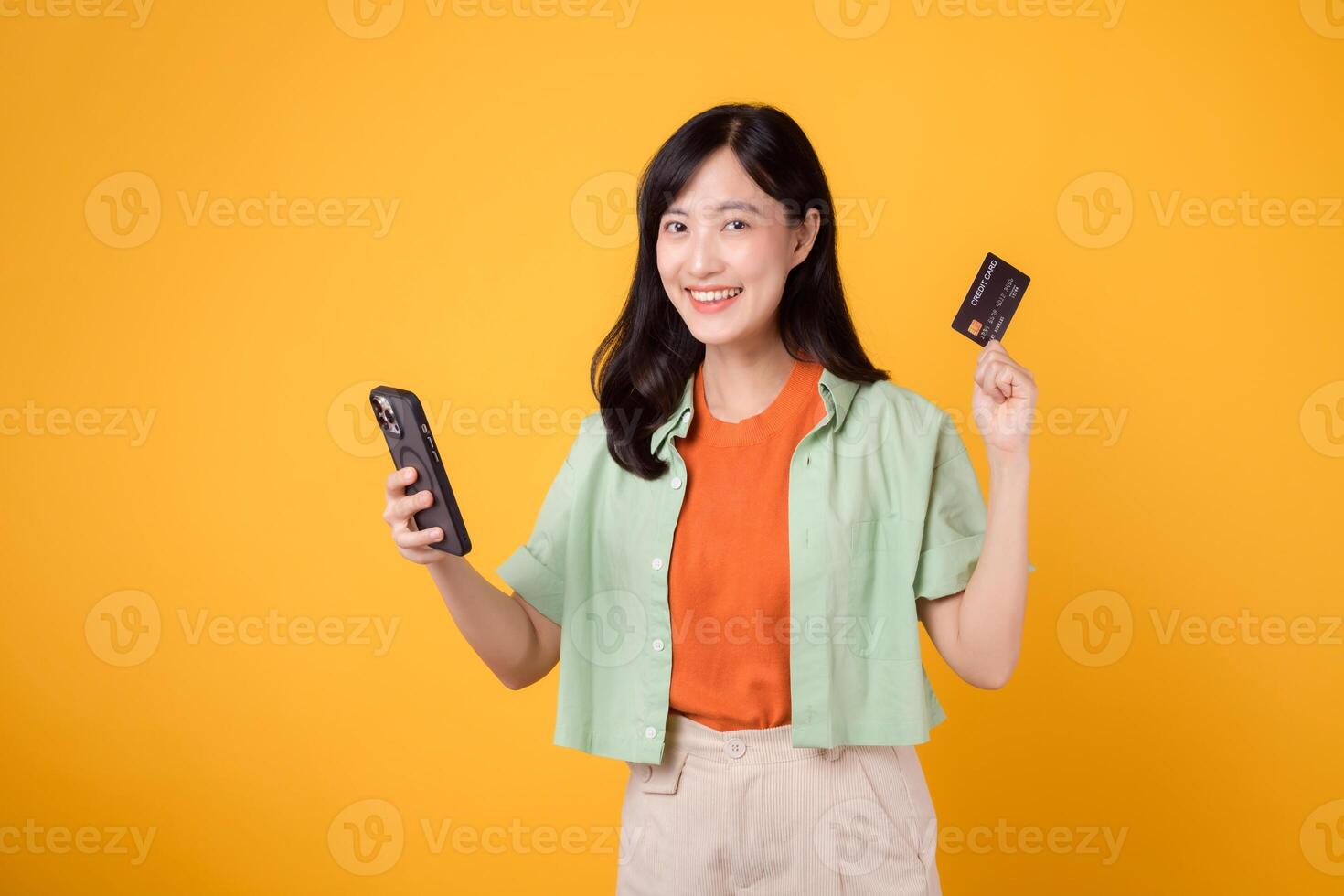 emoção do compras a partir de Móvel com uma cativante jovem ásia mulher dentro dela 30s, elegantemente vestido dentro laranja camisa e verde saltador, usando Smartphone para mostrar crédito cartão em amarelo estúdio fundo. foto