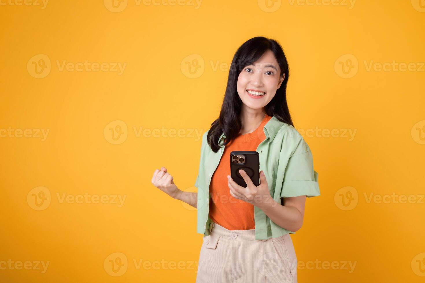animado jovem ásia mulher dentro dela 30s, vestido dentro laranja camisa e verde saltador, usando Smartphone com punho acima gesto em amarelo fundo. Móvel conceito com ótimo notícias. foto