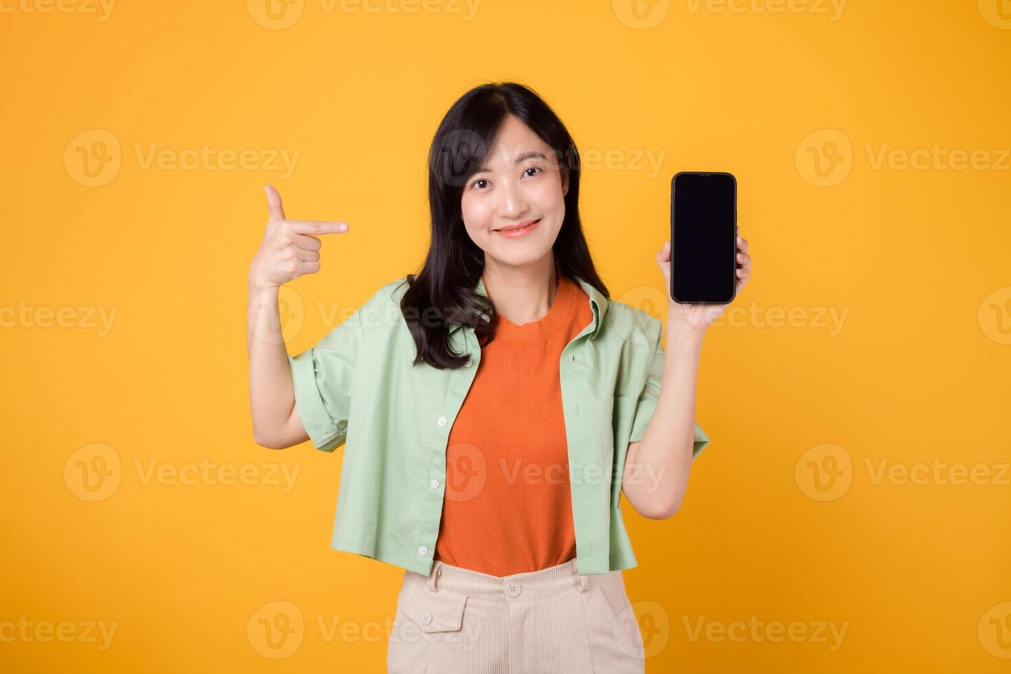 a inovação do Novo Móvel inscrição com jovem ásia mulher 30s, elegantemente vestido dentro laranja camisa e verde saltador, apresentando Smartphone tela com afirmativo gesto em amarelo fundo foto