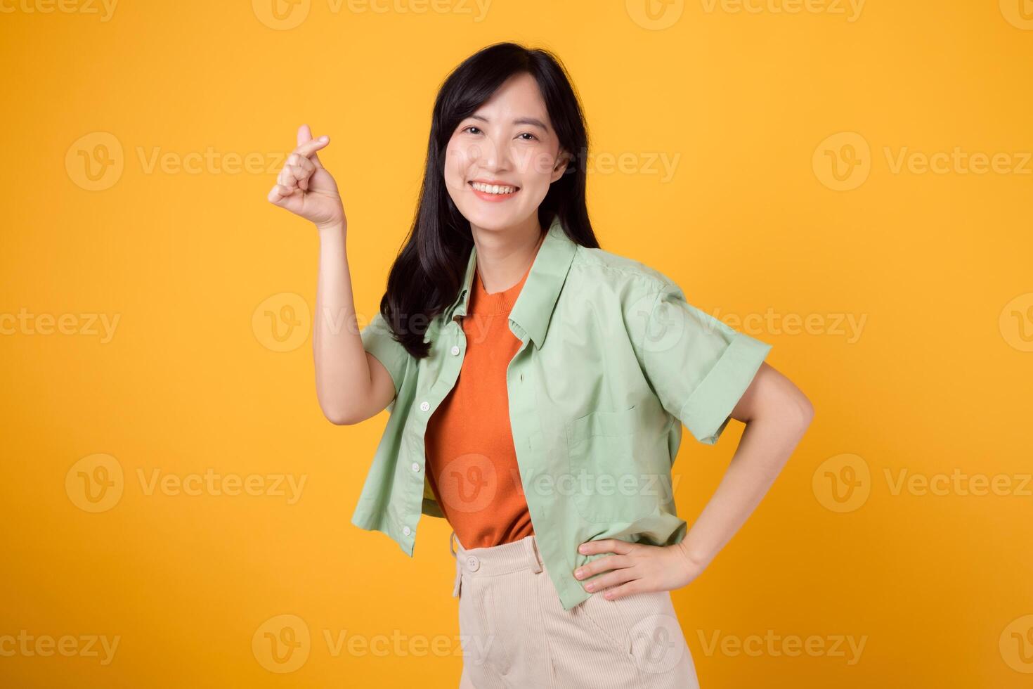 felicidade com uma jovem ásia mulher dentro dela 30s, vestido dentro a laranja camisa e verde saltador. dela mini coração gesto, mão em quadril, e □ Gentil sorrir transmitir uma profundo mensagem através corpo linguagem. foto